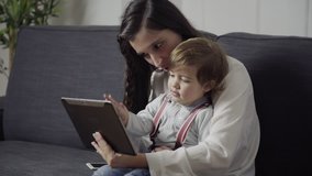 Smiling mother with cute toddler using digital tablet. Happy young woman sitting on couch with adorable little son and using tablet pc together. Wireless technology concept