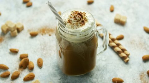 Glass jar with sweet caramel drink garnished with whipped cream and served with straw on table among nuts