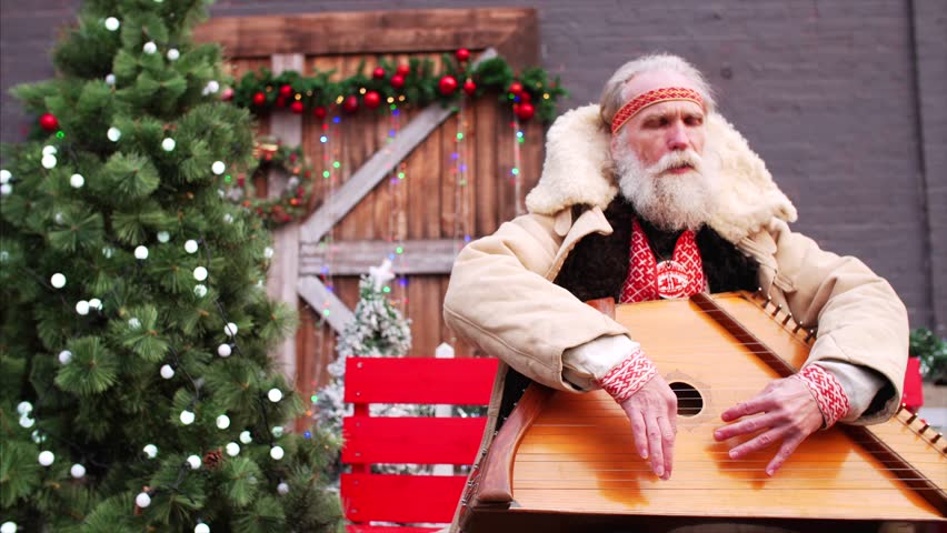 Sliding portrait of old man with white beard in rustic national russian costume sitting on the bench near Christmas trees, playing dulcimer and singing very emotionally