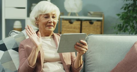 Caucasian senior grey haired woman having a videochat on the tablet device via webcam in the nice living room. Indoor.isolated, covid-19, quarantined, concept, isolation, home, protection, coronavirus