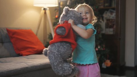 Happy baby girl playing and dancing with soft toy Teddy bear home. Portrait of expressive little girl hugging her plush bear friend, indoor shot. Little girl playing with teddy bear
