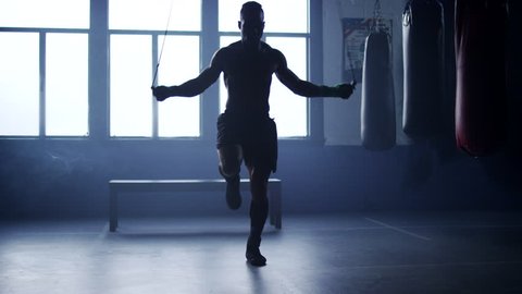 A shirtless, muscular African American athlete skillfully jump ropes in an old, hazy boxing gym