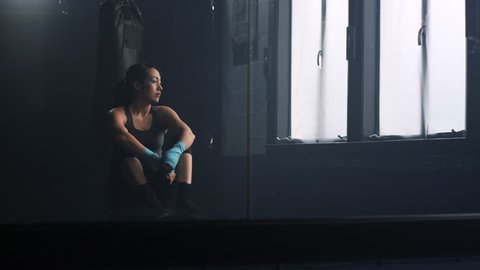 An attractive, female athlete sits on a bench in a boxing gym, reflected in a mirror