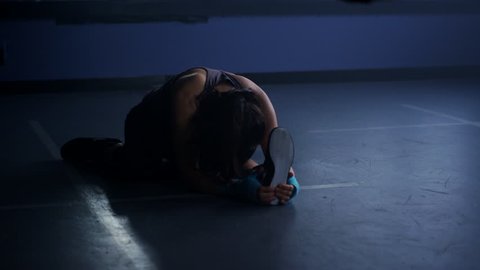 A young, female athlete warms up with hamstring stretches in a dimly lit boxing gym