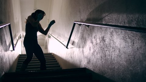 A muscular female fighter shadow boxes in a dark stairwell. She turns towards the camera and shakes out her muscles.