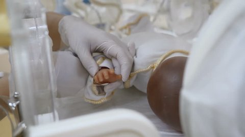 Newborn in maternity clinic concept. Close up of doctor hands setting finger sensors to infant palm in incubation apparatus or couveuse. A doll in incubator as if a real infant or premature kid. Child