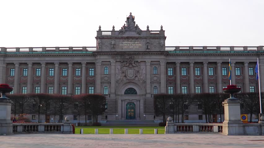Front of the Swedish Parliament House in Stockholm, Sweden. Green grass, trees and ornaments in front of the building. Bright, cloudy sky in the background.