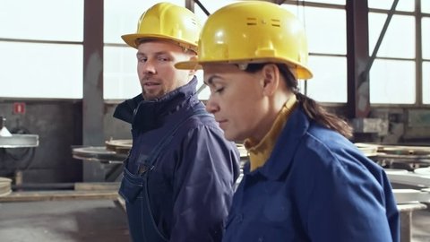 Tracking shot of male and female workers in overalls and hard hats walking through metal fabrication facility and talking