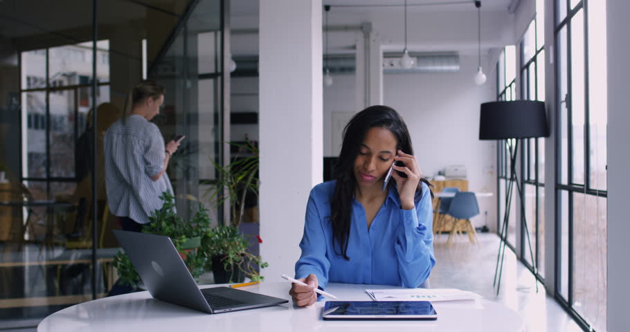 Dark skinned female employee talking on cellphone and checking information with stylus pencil on touch pad while her coworking colleague walking on background, Slow motion effect on 4K RED camera | Shutterstock HD Video #1022539297