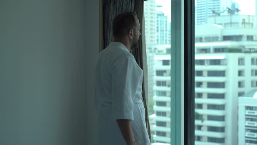 Handsome man in bathrobe standing by the window and admires the view 
