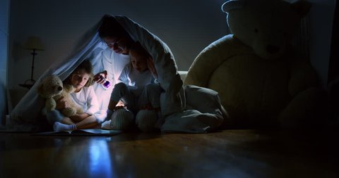 Slow motion of young mother is reading a goodnight story to her little daughters in the dark illuminating with a torch under the blanket. Concept of family, childhood, love, health, education