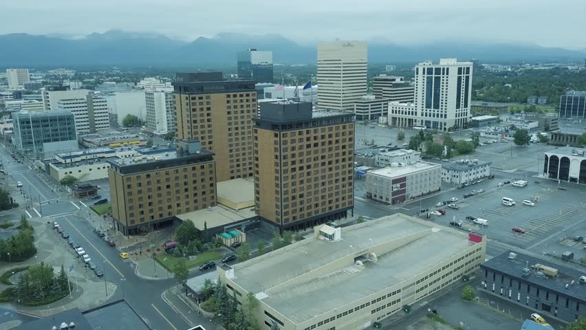 Aerial drone panning above the city of Anchorage, Alaska USA with high rises and buildings surrounded by vegetation with large mountains in the foggy distance