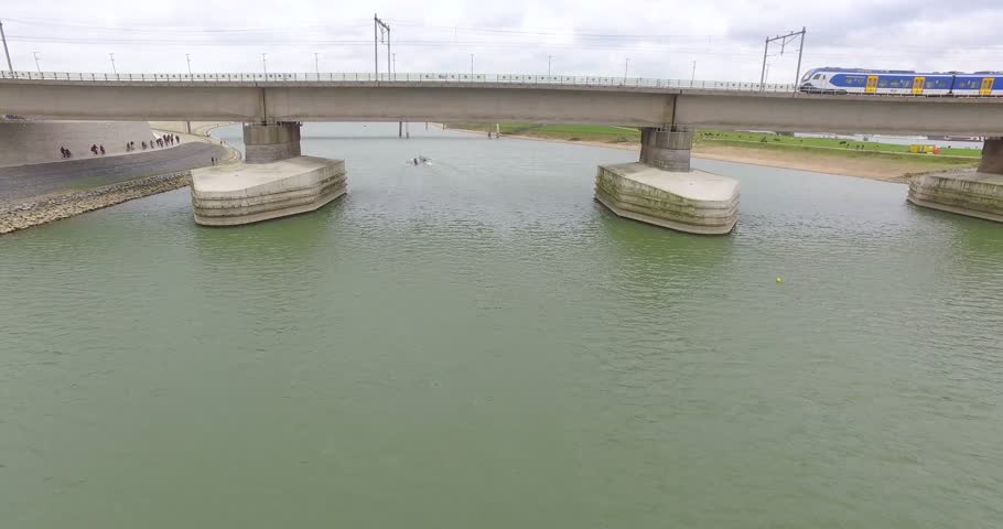 Areal footage above a river near a massive bridge on which a train runs through the frame.
