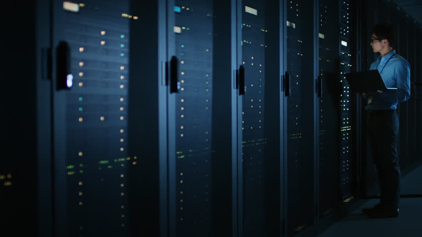 In Dark Data Center: Male IT Specialist Stands Beside the Row of Operational Server Racks, Uses Laptop for Maintenance. Concept for Cloud Computing, Artificial Intelligence, Supercomputer - Powered by Shutterstock - Get 15% off with code: PIKWIZARD15