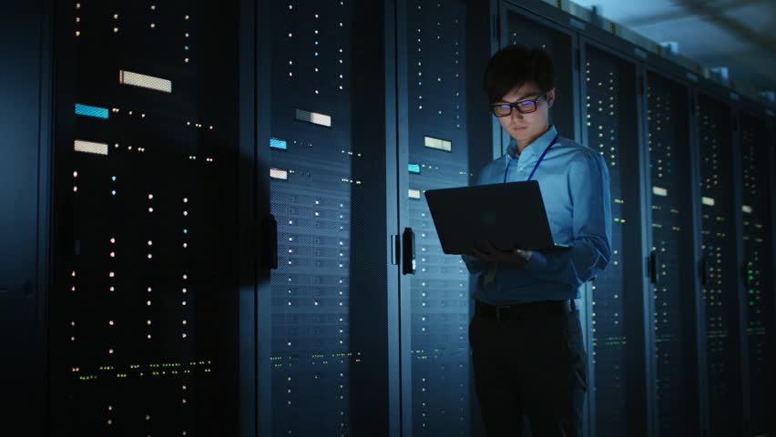 In Dark Data Center: Male IT Specialist Walks along the Row of Operational Server Racks, Uses Laptop for Maintenance. Concept for Cloud Computing, Artificial Intelligence, Supercomputer, Cybersecurity Royalty-Free Stock Footage #1022552605
