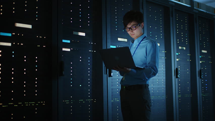 In Dark Data Center: Male IT Specialist Walks along the Row of Operational Server Racks, Uses Laptop for Maintenance. Concept for Cloud Computing, Artificial Intelligence, Supercomputer, Cybersecurity - Powered by Shutterstock - Get 15% off with code: PIKWIZARD15