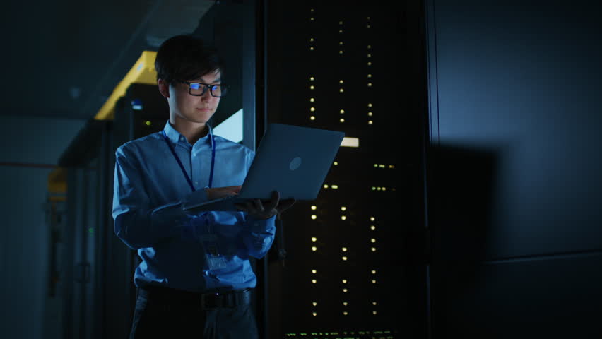 In Dark Data Center: Male IT Specialist Stands Beside the Row of Operational Server Racks, Uses Laptop for Maintenance. Concept for Cloud Computing, Artificial Intelligence, Supercomputer. 8K RED Royalty-Free Stock Footage #1022552608