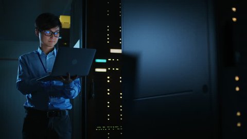 In Dark Data Center: Male IT Specialist Stands Beside the Row of Operational Server Racks, Uses Laptop for Maintenance. Concept for Cloud Computing, Artificial Intelligence, Supercomputer. 8K RED
