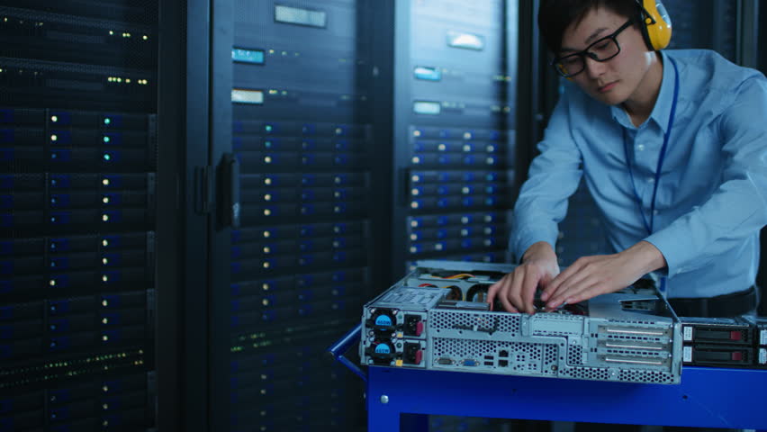 In the Modern Data Center: IT Technician Wearing Protective Headphones Working with Server Racks, on a Pushcart Installing New Hardware. Engineer Doing Maintenance and Diagnostics of the Database. Royalty-Free Stock Footage #1022552611