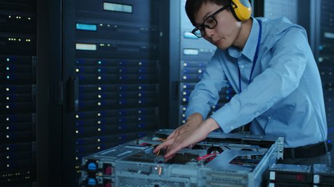 In the Modern Data Center: IT Technician Wearing Protective Headphones Working with Server Racks, on a Pushcart Installing New Hardware. Engineer Doing Maintenance and Diagnostics of the Database.
