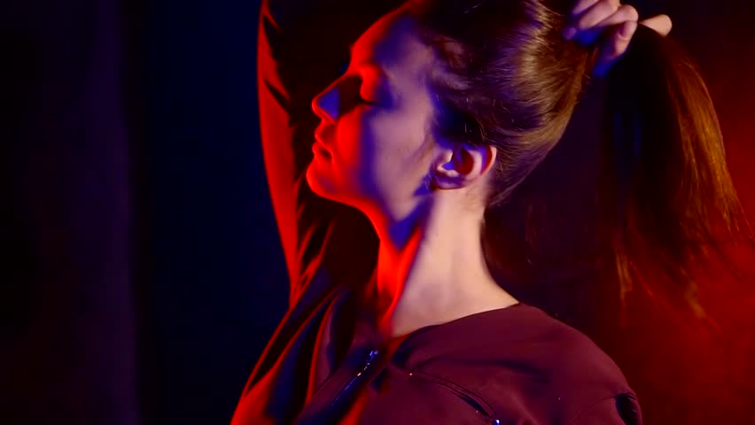 relaxed young woman is standing alone in dark room, holding her hair up by hands
