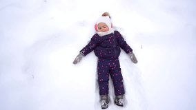 Cute sweet little girl playing outdoors in winter snowy park. Top view full length portrait of happy smiling little baby laying on her back in fresh fluffy snow on ground. Slow motion video footage.
