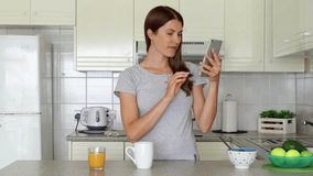 Young woman in pajamas enjoying early morning standing on kitchen drinking coffee or tea. Sleepy female housewife using mobile phone talking via messenger app having video chat