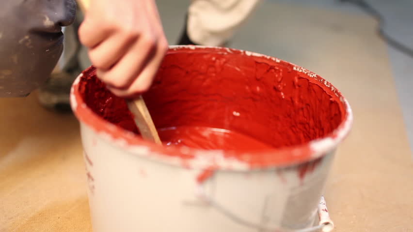 Close up of a painter stirring with a wooden stick in a paint bucket full of red paint.