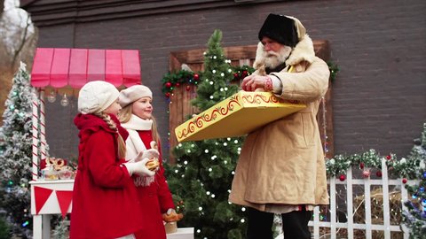 Whitebearded man is showing cracknels for two cut girls in red coats. Christmas fair, beautiful decorations
