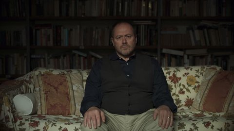 Portrait of a serious Italian man sitting on a couch with bookshelves filled with books in the background with soft natural lighting. Medium shot on 4k RED camera.