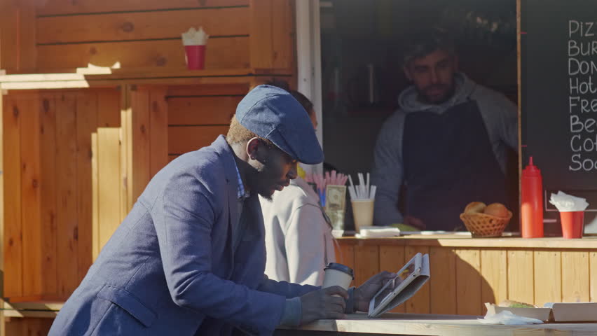 Cross section shot of young African American man drinking coffee near food booth and checks social media on tablet computer while Caucasian brunette woman buying coffee on background plane