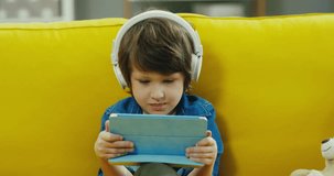 Close up of the small cute boy inthe big white headphones sitting on the yellow couch and watching cartoons on the tablet computer. Indoor.