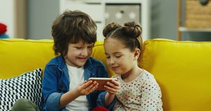 Close up of the Caucasian small and cute sister and brother sitting on the yellow couch in the living room and watching something or playing on the smartphone device. Indoors.