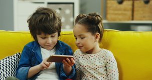 Two little kids - boy and girl - sitting on the yellow sofa and watching something or playing on the smartphone. Indoor.
