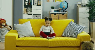 Caucasian little pretty girl sitting on the yellow sofa in the nice living room and using tablet device.
