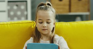 Close up of the little cute girl with fair hair playing on the tablet device while sitting on the yellow couch at home and looking at the side.