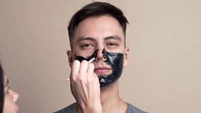 Pretty young guy in applies black face cosmetic mask in slow motion, video portrait, face care, selfie video, beauty blogger, face cosmetics