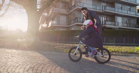 Daughter child girl learning riding bycicle with dad teaching in city.Growing,childhood,active safety family.Sidewalk urban outdoor.Warm sunset cold weather backlight.4k slow motion 60p side video