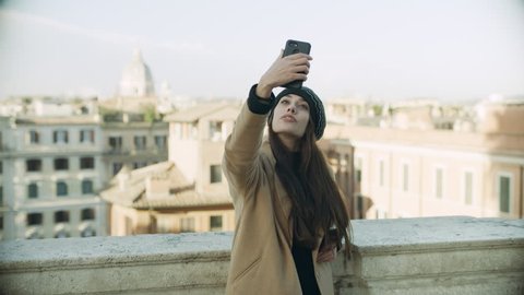 Fashionable Italian woman modeling taking selfies with a mobile phone on  balcony overlooking buildings and churches of Rome, with soft day lighting. Wide shot on 4k RED camera.