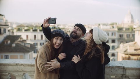 Group of Italian friends taking selfies with a phone on a balcony overlooking buildings and churches of Rome, with soft day lighting. Wide shot on 8k helium RED camera