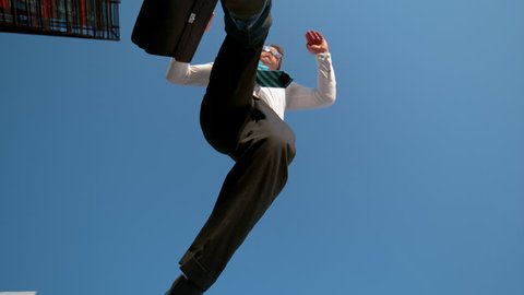 SLOW MOTION, BOTTOM UP: Cheerful young businessman jumps over the camera after getting promoted. Happy young Caucasian man carrying a briefcase jumping in the air after work on a sunny spring day.