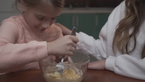 Little girl mixes eggs with flour in bowl sitting at the table in the kitchen with her older sister. Elder girl praises her sister