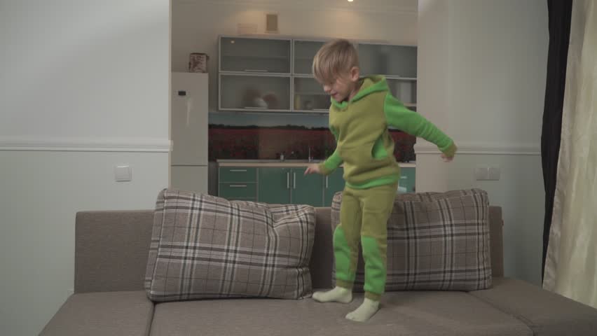 Cute jouful boy in green pajamas jumps on the sofa at home. Child grabs pillow and throws in on the floor. Happy cheerful kid. Slow motion.
