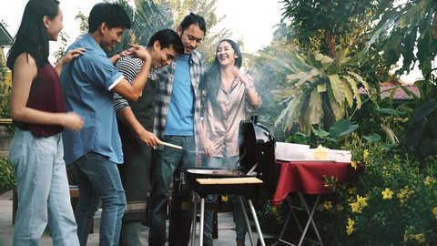 Friends trying to cook raw burger in bbq grill outdoor. Group of asian, caucasian young man and woman standing around bbq grill, laughing and having fun together with burn meat. House party concept.