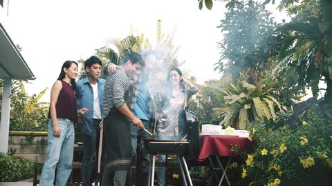 Friends trying to cook raw burger in bbq grill outdoor. Group of asian, caucasian young man and woman standing around bbq grill, laughing and having fun together. House party concept.