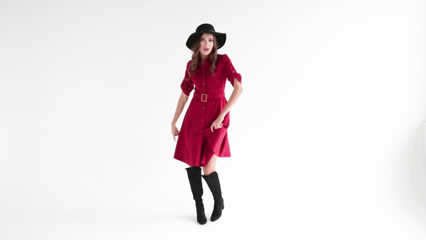 Young funny girl in beautiful red dress is dancing and laughing. A girl with blond hair is dancing on a white background. A girl is dancing in a red dress and black hat. The girl is spinning and