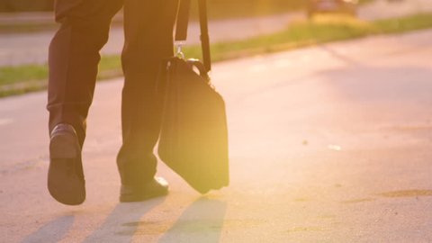 SLOW MOTION, LENS FLARE, CLOSE UP: Pessimistic young man slowly walking home after an unsuccessful job interview. Businessman lost his job and walks home with his briefcase in hand at golden sunset