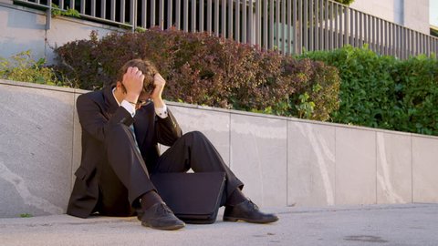 SLOW MOTION: Sad man in a suit sits on the sidewalk and buries his head in his hands after a stressful day at the office. Young Caucasian businessman grieving after losing his job and going bankrupt.