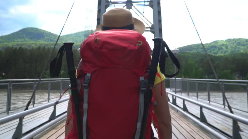 The woman the traveler with a red backpack goes along the cable-stayed bridge through the mountain river.