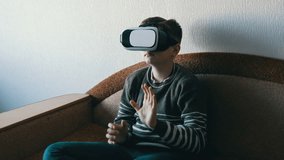 Teen boy in white glasses of virtual reality or vr on his head sitting on the couch in the room and playing